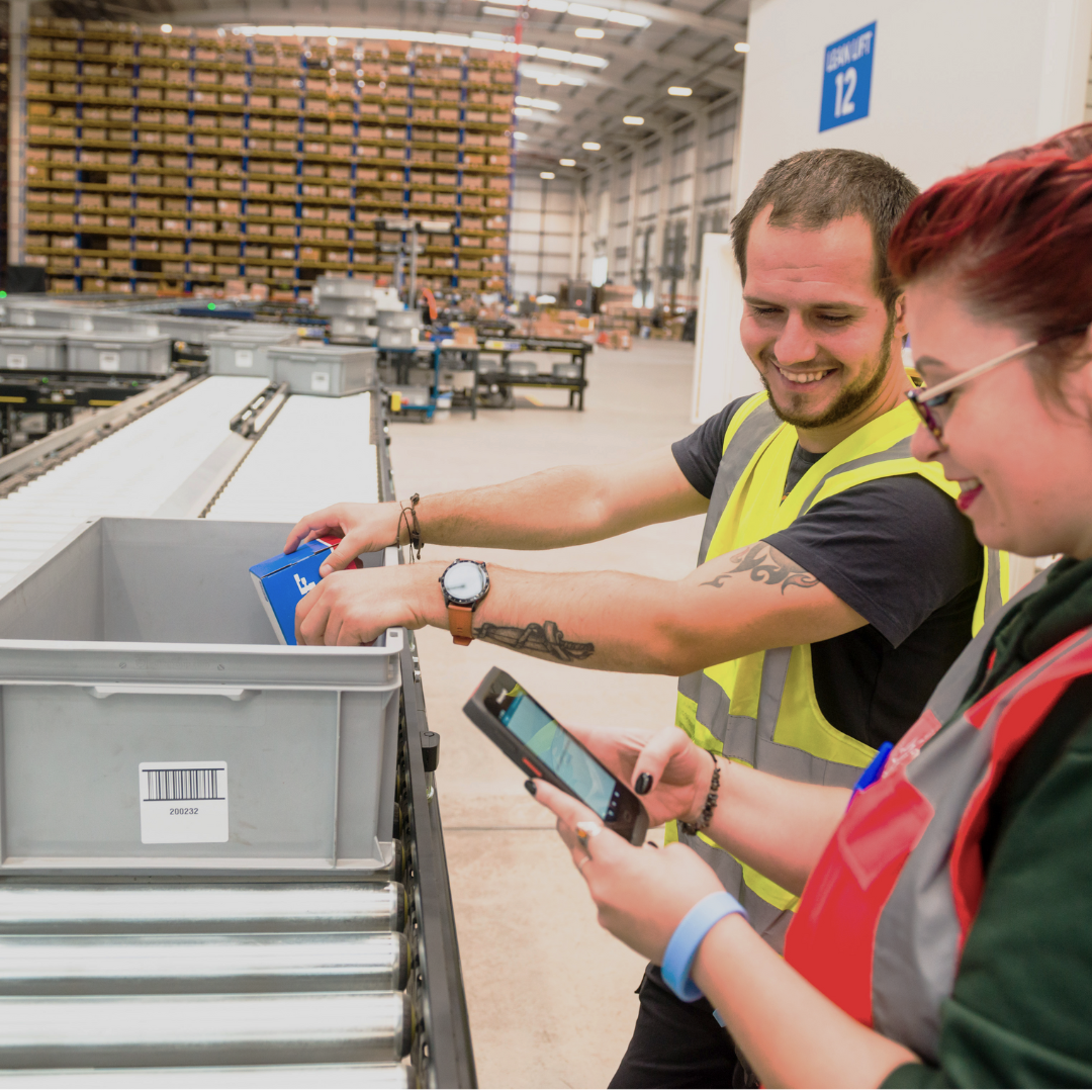 A woman and a male, on the sorting line in a warehouse, checking orders collected in a grey bin on a conveyor, both in PPE