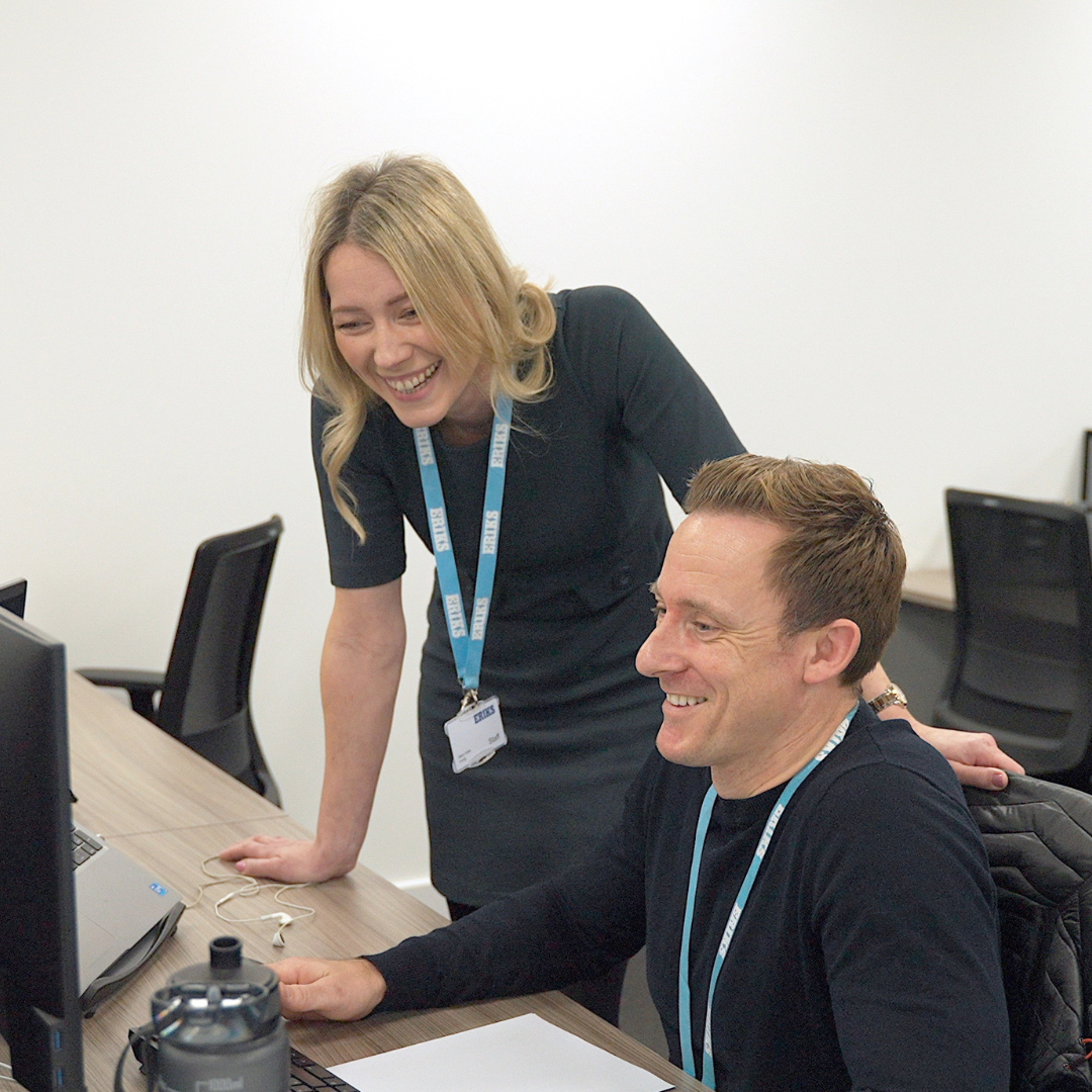 A woman standing looking at a male colleagues screen, both smiling in an office setting