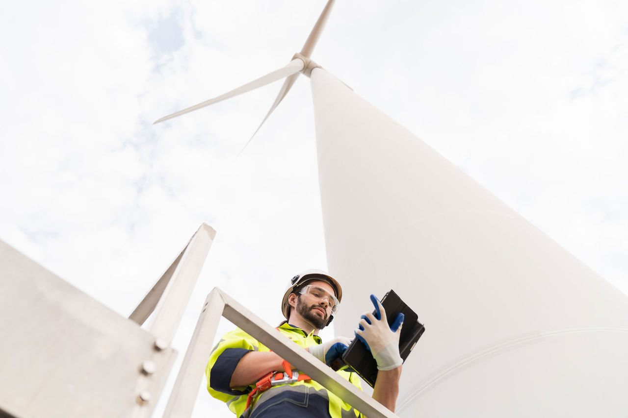 Male engineer working with plan inspecting or maintenance of wind turbines at windmill field farm. Male engineer using computer tablet control or monitoring wind turbine system at wind turbines farm