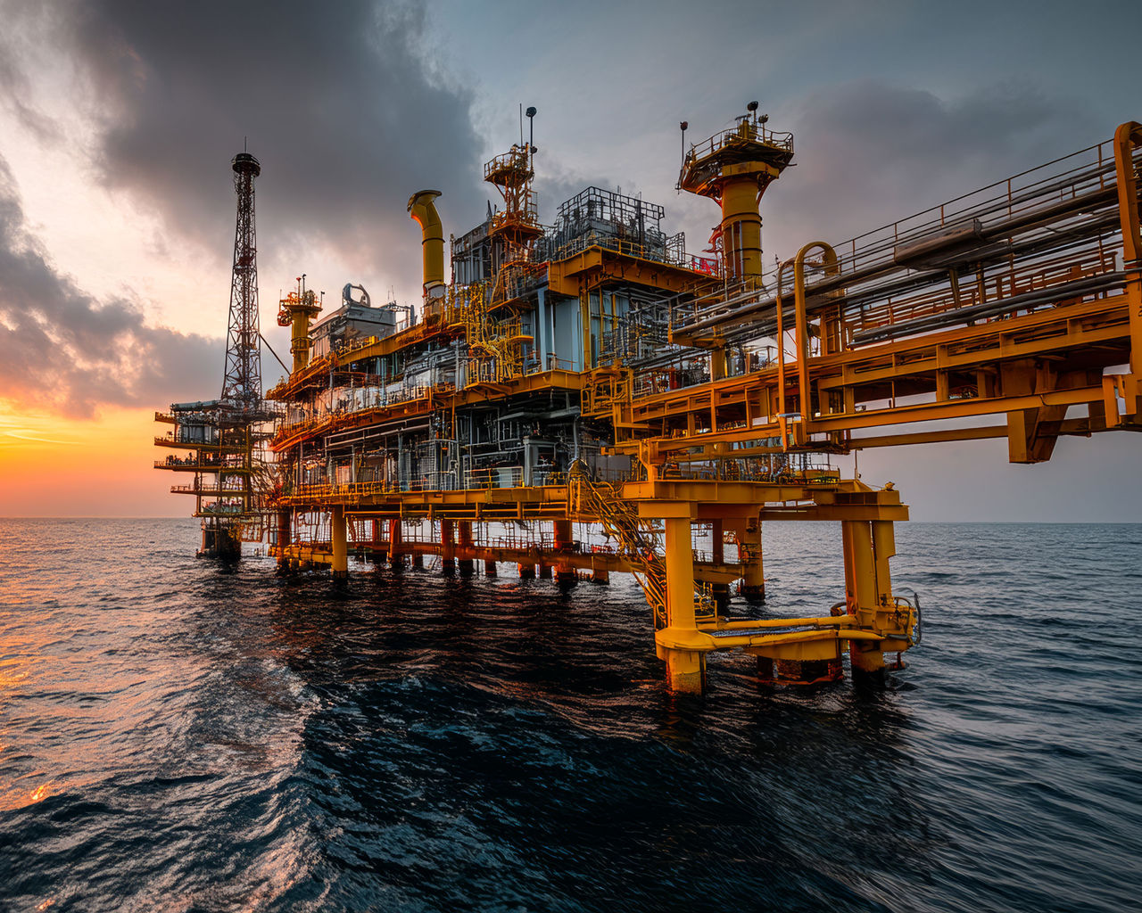 Oil rig at sunset over calm waters. An oil rig stands tall above the ocean, silhouetted against a vibrant sunset with clouds and shimmering water.