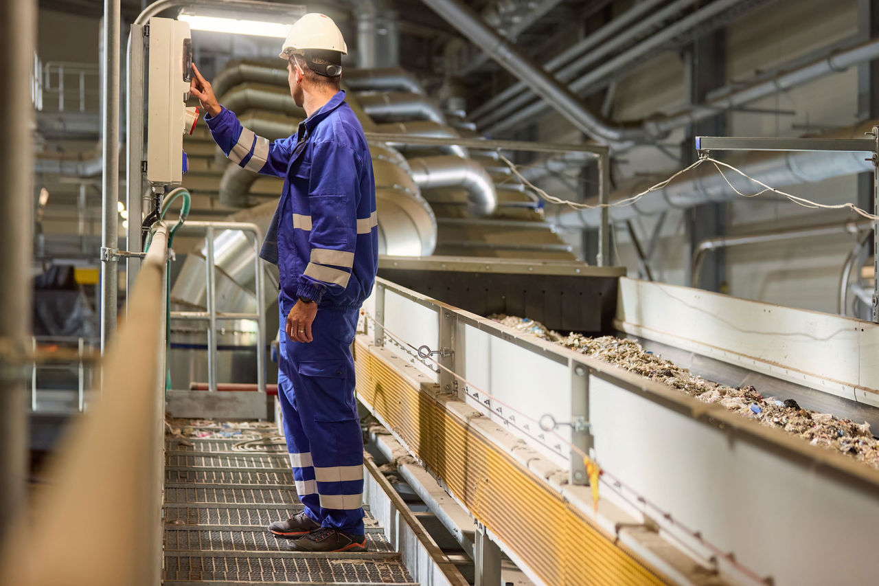 A technician wearing blue overalls and a white helmet operates machinery at an industrial recycling facility where he manages the sorting and processing of refuse-derived fuel (RDF) on a conveyor belt
