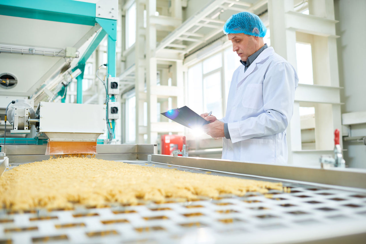 Person in overcoat and hairnet standing in front of food production line writing on clipboard