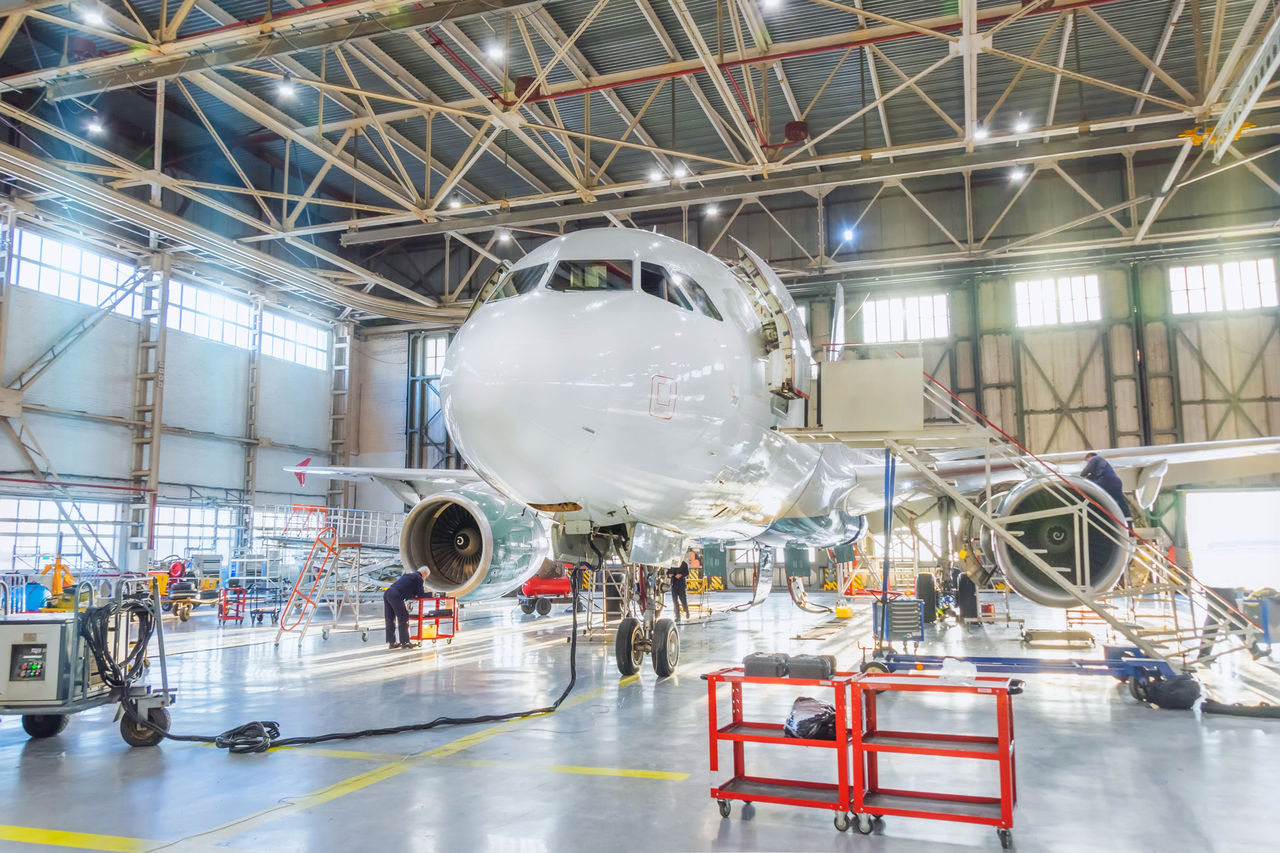 Aircraft inside the aviation hangar, maintenance service. Airplane technician worker working around. Bright light outside the gate. Aircraft inside the aviation hangar, maintenance service. Airplane technician worker working around. Bright light outside the gate.