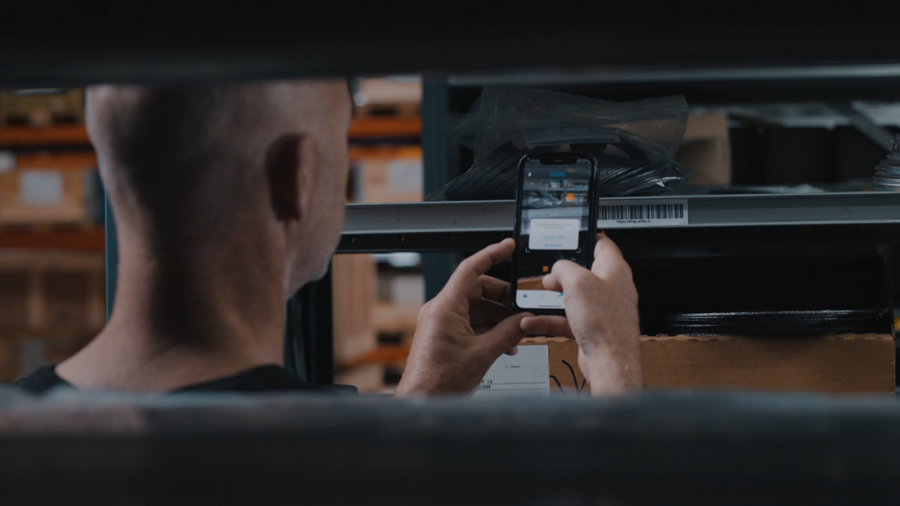 Warehouse setting with a person's hand holding a phone scanning a barcode label on a shelf holding products